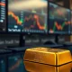 A gold bullion bar on a desk with financial market charts in the background.