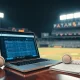 A laptop showing a prediction market next to a baseball and scorebook in a stadium press box.