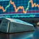 A silver bullion bar on a desk with a financial chart showing a price decline in the background.