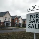A 'For Sale' sign in front of a UK housing development under cloudy skies.
