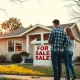 A couple looks at a for sale sign in front of a house, representing first-time homebuyer opportunities.