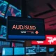 AUD/USD trading screen showing a decline from recent highs on a dimly lit trading floor.