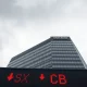 A somber view of a major Australian bank headquarters with a red stock ticker showing falling share prices, reflecting market pressure from budget changes.