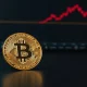 Bitcoin coin on dark surface with red downward trend line in background, representing price correction analysis.