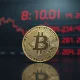 A cracked Bitcoin coin on a reflective surface with a blurred red stock market ticker in the background.