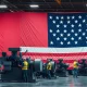 American flag draped over industrial machinery in a busy factory with workers operating equipment