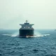 Oil tanker navigating the Strait of Hormuz under overcast skies with a military vessel in the distance.