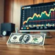 U.S. dollar bill on desk with stock market chart on monitor in background