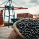 Burlap sack of coffee beans at a port with cargo ship in background, representing global coffee trade and supply chain factors.