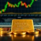 Gold bar and coins on a desk with a financial chart in the background, representing precious metals market analysis.