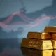 Stacked gold bars with blurred map of Strait of Hormuz in background, representing safe-haven demand amid geopolitical tensions.