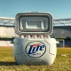 Miller Lite limited-edition Matchball cooler shaped like a soccer ball, open and holding a 12-pack of beer on a grassy field near a stadium.
