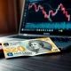 New Zealand Dollar banknote on desk with laptop showing a downward forex chart