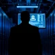 Silhouette of a person facing away from camera in a server room, with a glowing monitor showing anonymous user data.
