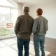 Couple standing in empty living room looking at For Sale sign through window, contemplating selling price