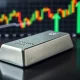 Silver bullion bar on dark wood with financial charts in background