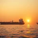 Oil tanker near the Strait of Hormuz with a naval warship in the background during a geopolitical crisis.