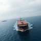 Oil tanker navigating the Strait of Hormuz under overcast skies, with naval presence in the distance.