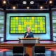 A television studio set for the Wordle game show with a large puzzle screen and host Savannah Guthrie at the podium.