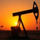 Oil pumpjack silhouette against sunset sky with faint US and Iran flags
