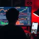 Silhouette of a teenager in a dark room using a computer with cryptocurrency charts and a smartphone displaying Instagram, representing a crypto theft investigation.