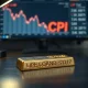 Gold bar on desk with a blurred financial monitor showing a downward chart and CPI label