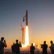 SpaceX rocket launching at dawn with reporters and analysts in foreground.