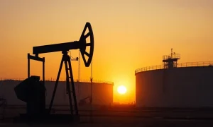 Oil pumpjack silhouetted against a sunset sky with storage tanks in the background