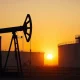 Oil pumpjack silhouetted against a sunset sky with storage tanks in the background