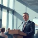 FCA official speaking at a podium in a London conference room about AI regulation in financial services.