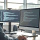 Developer working at a dual-monitor setup with code on screens in a bright office
