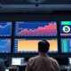 Traders watching exchange screens showing falling stock charts, rising oil and a bitcoin ticker