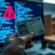 Hand holding a driver's license in front of a computer screen with code, representing the IDScan data breach
