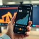 Financial chart on a smartphone showing an upward trend, with oil and dollar imagery in the background.