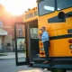 Yellow school bus parked outside a school as an administrator stands by the open door